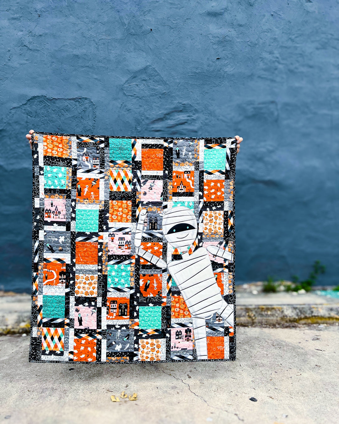 A Halloween themed Dashing Duo Quilt with mummy popping in on the right side of quilt held in front of a gray wall