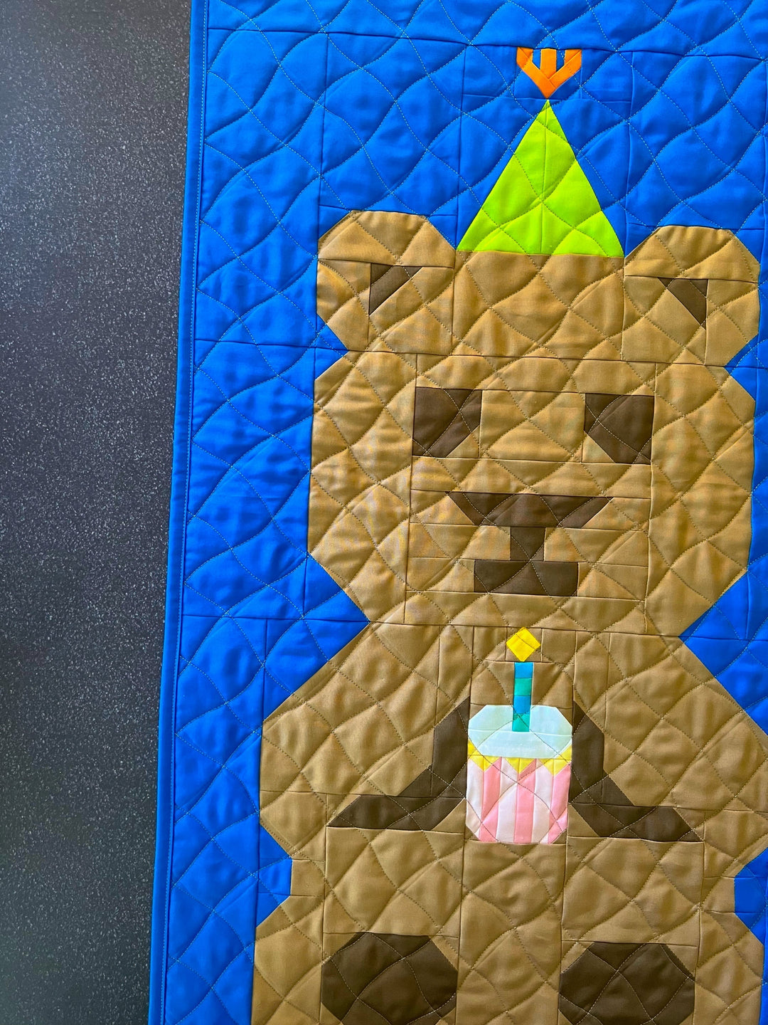 A close-up of a quilt featuring a stylized brown bear holding a small birthday cake against a bright blue background, with a dark textured surface visible along the left edge.