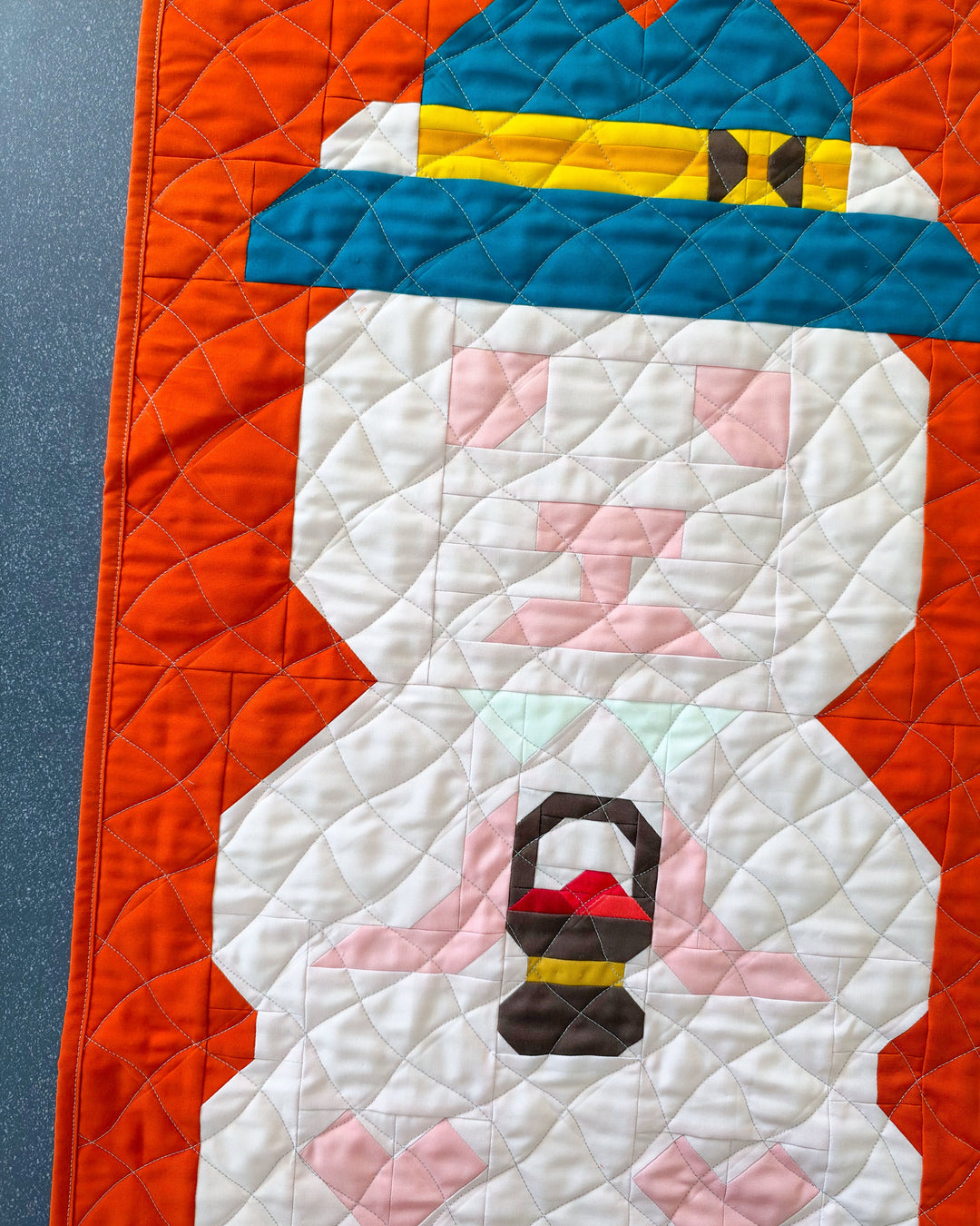 A close-up of a quilted fabric featuring a stylized autumn themed bear design with a bright orange background, pale pink body, pink facial details, a blue hat, and a small brown-and-red cherry basket motif.