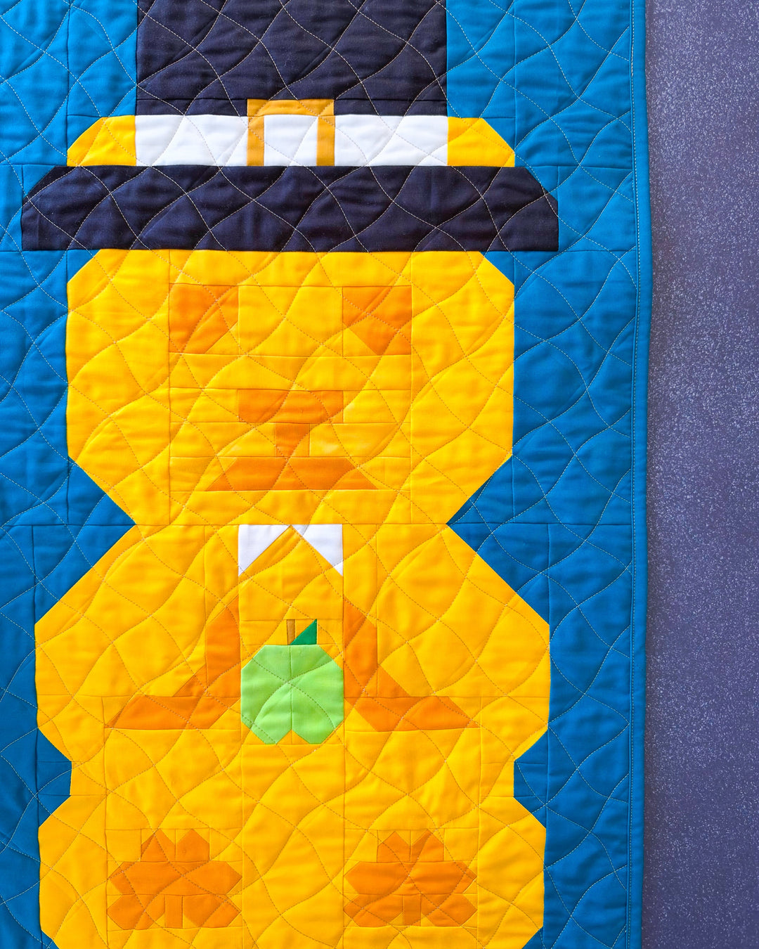 A close-up of a quilted textile featuring a stylized pilgrim bear figure in yellow with a black hat, white band, and green apple, set against a blue background.