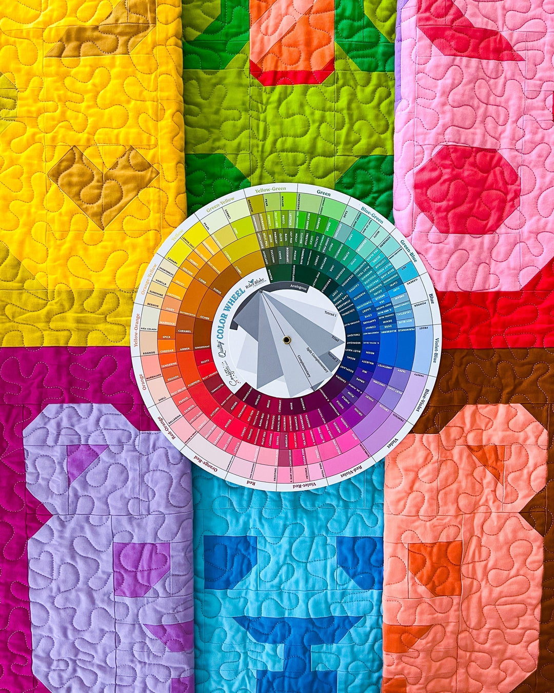 A circular color wheel labeled 'Confetti Cotton Quilty Color Wheel Riley Blake Designs' is centered over a colorful quilted made of sewing themed bear blocks in yellow, green, pink, purple, blue, orange, and brown.