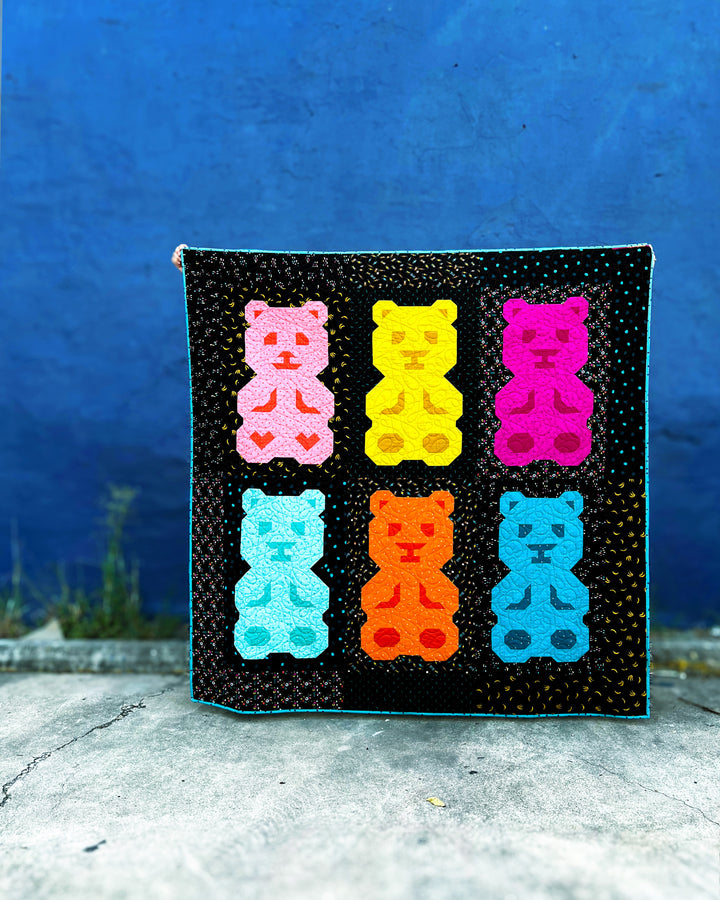 The Beary Bunch quilt is colorful gummy bear-shaped quilt blocks with a black background against a blue wall and concrete floor in the backdrop.