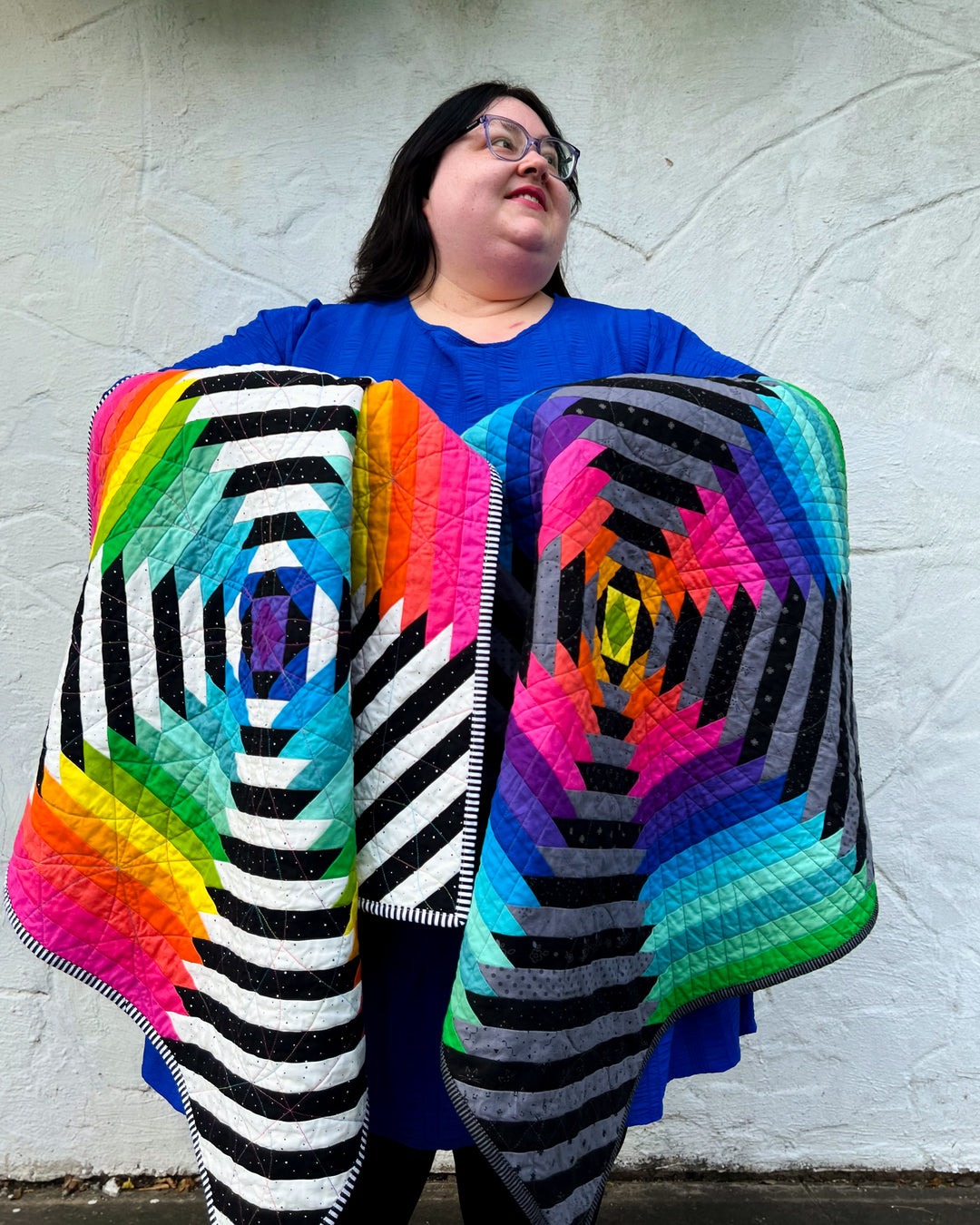 Person holding two colorful Supercolossal quilts draped over their arms against a white wall.