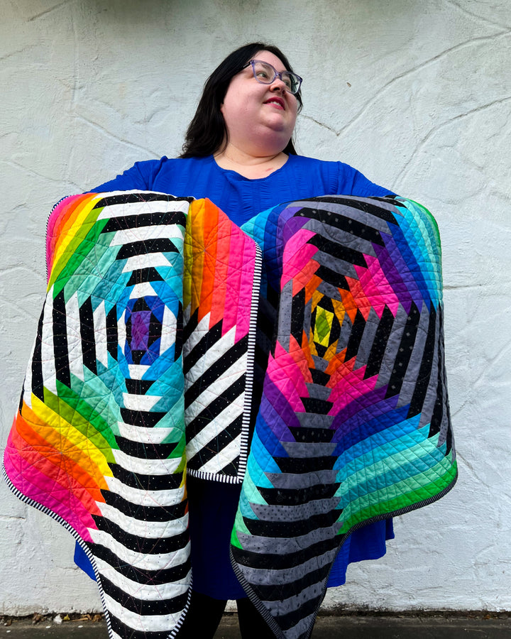 Person holding two colorful Supercolossal quilts draped over their arms against a white wall.