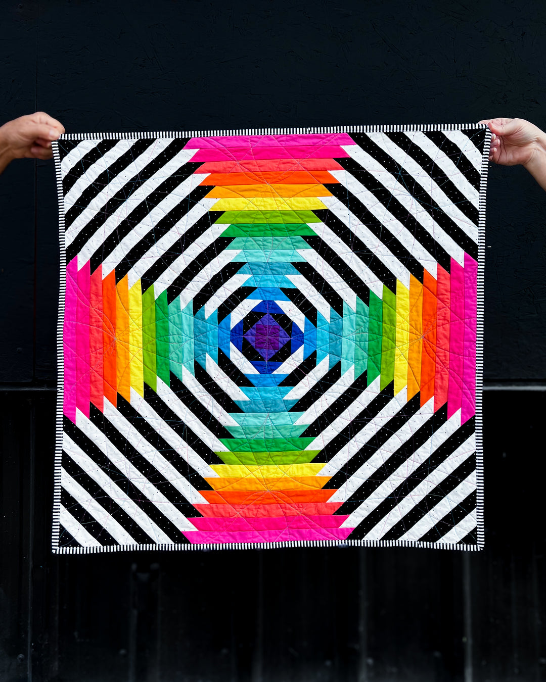 Supercolossal quilt with geometric pattern held by two hands against a black background