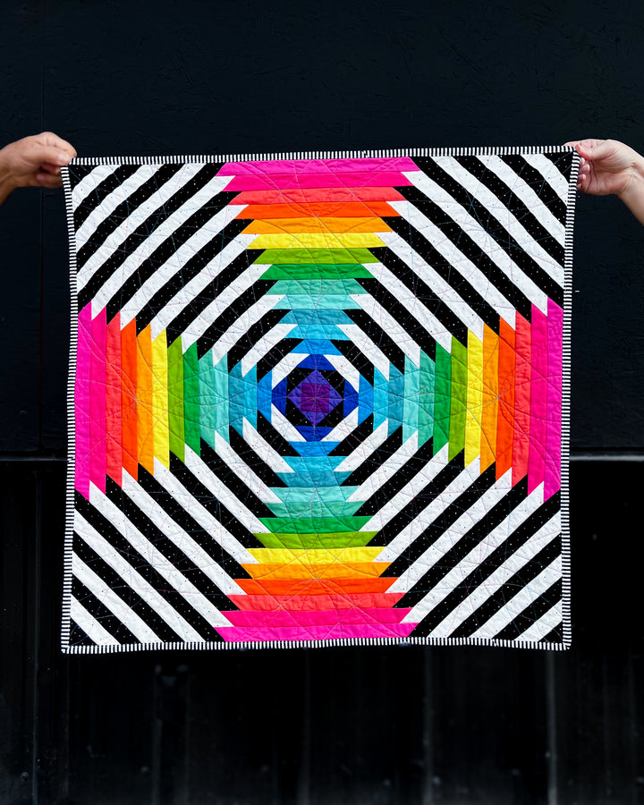 Supercolossal quilt with geometric pattern held by two hands against a black background