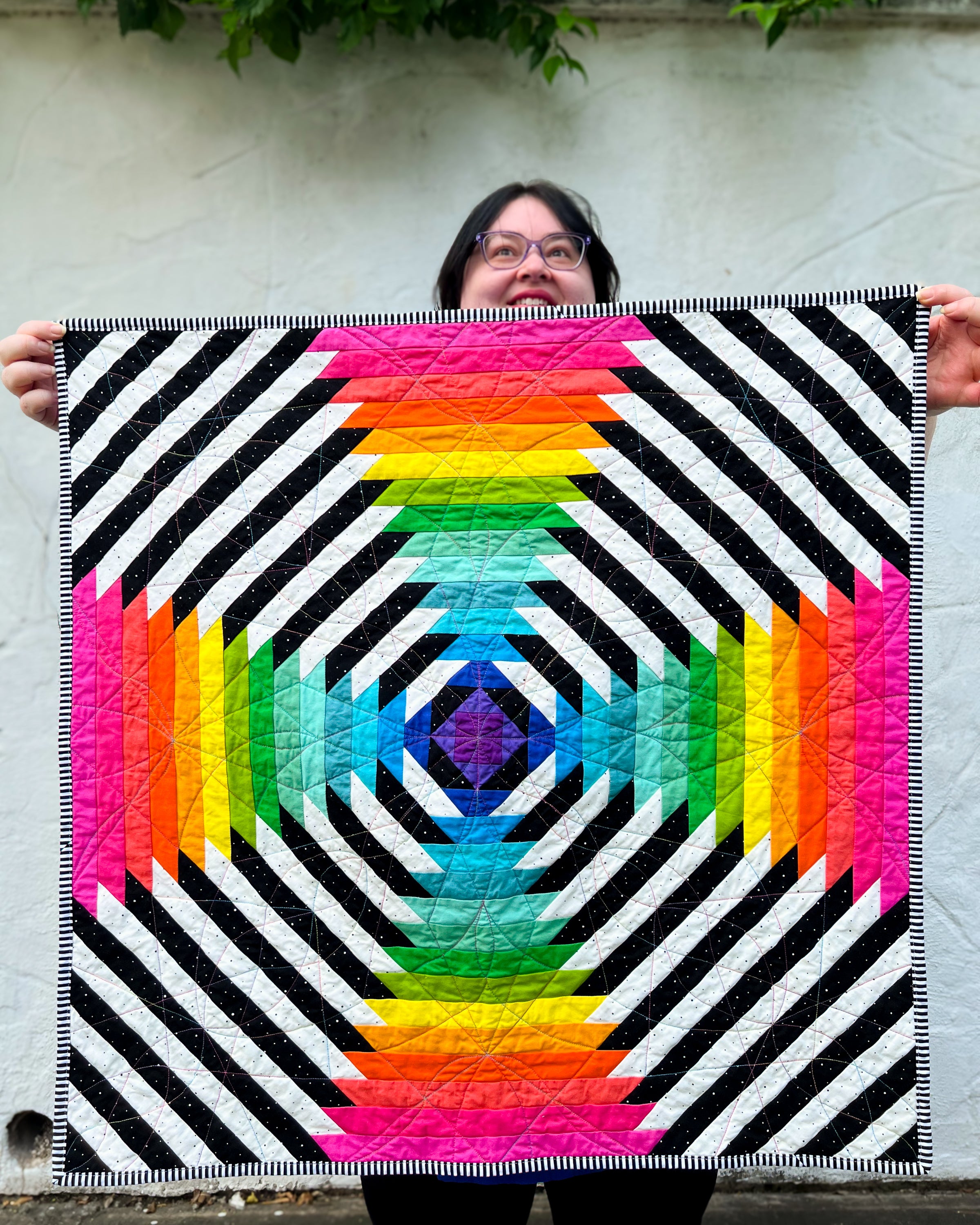 Person holding a colorful Supercolossal pineapple quilt with geometric pattern against a light-colored wall