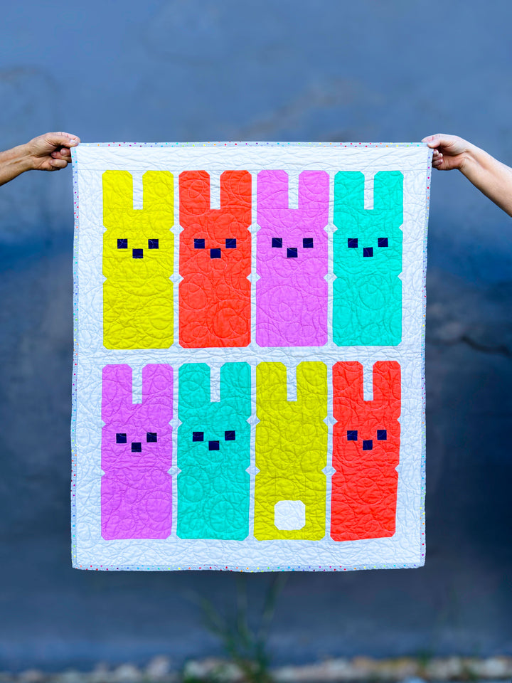 Hoppy Bunnies a colorful quilt with bunny patterns held by two hands against a blue background
