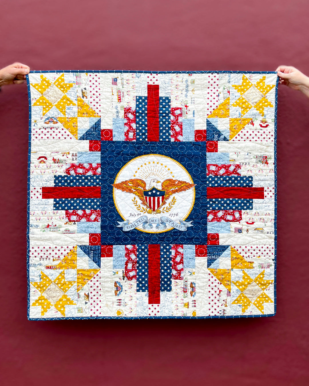 Converging Paths Quilt with a central emblem panel from the RBD Independence Collection being held by two hands against a red background