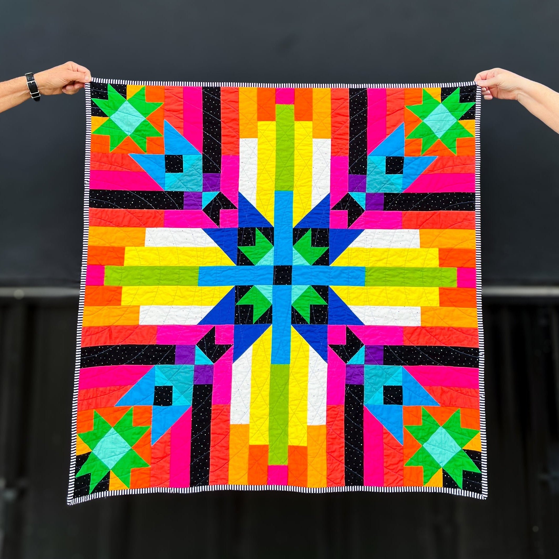 Converging Paths, a colorful geometric quilt held by two hands against a dark background