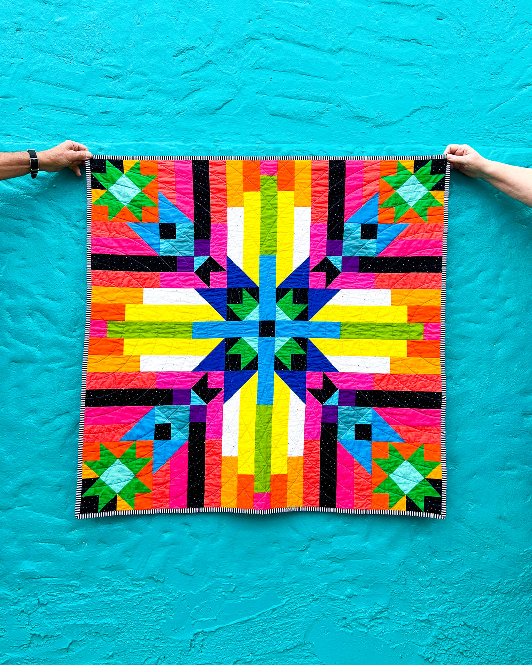 Converging Paths, a colorful geometric quilt held up against a blue background