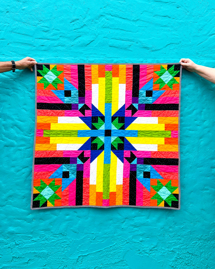 Converging Paths, a colorful geometric quilt held up against a blue background