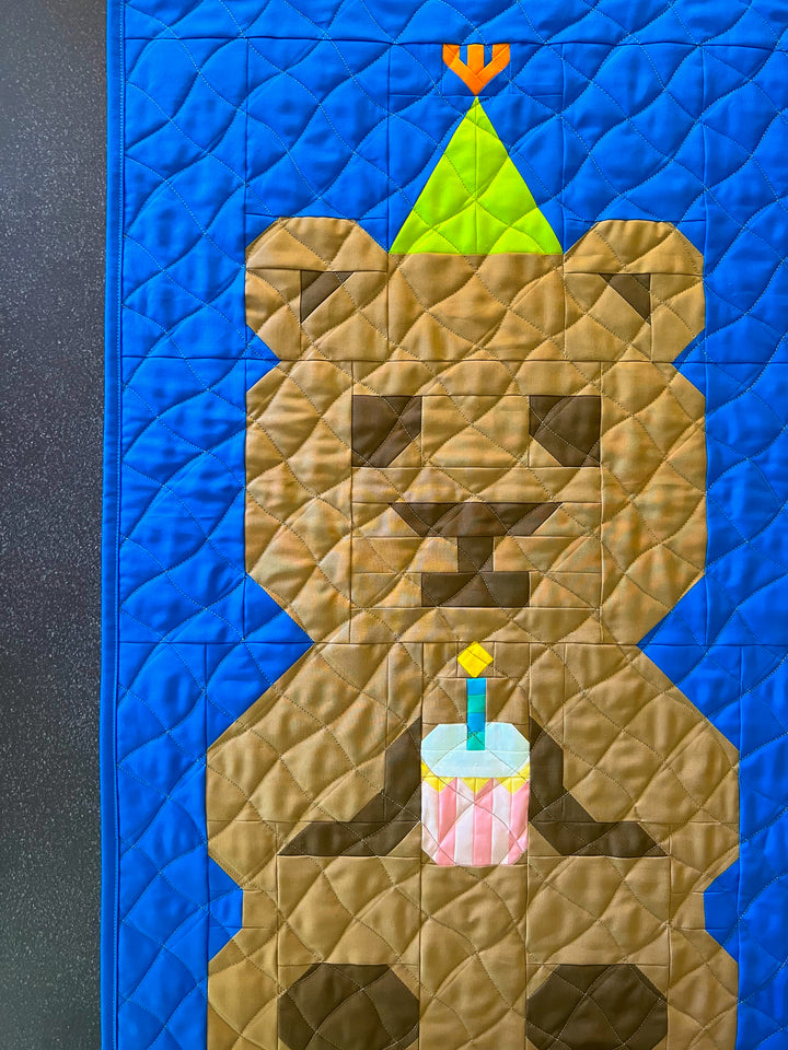 A close-up of a quilt featuring a stylized brown bear holding a small pink and white cupcake, with a bright blue quilted background and a lime-green party hat on the bear&