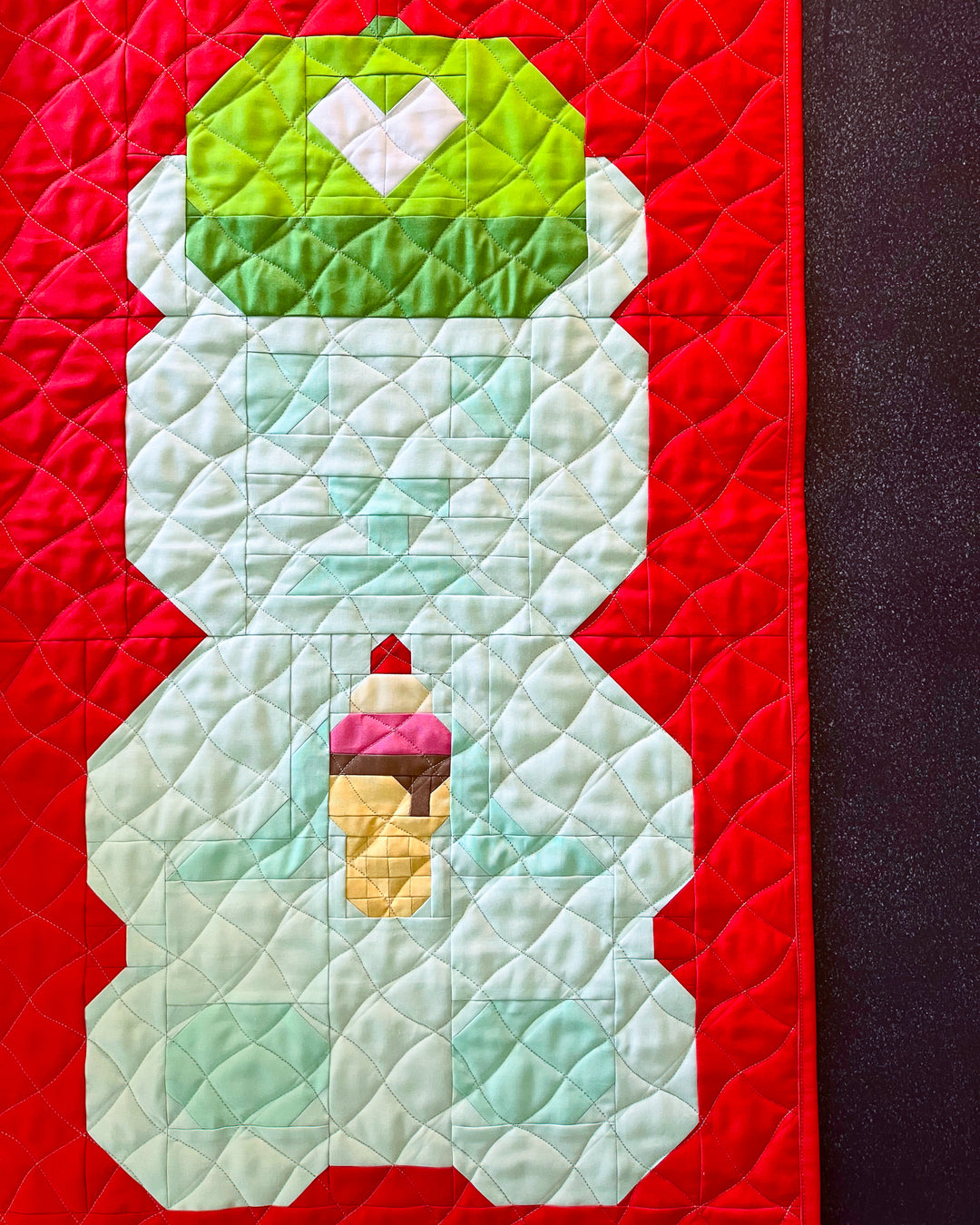 A close-up of a quilt featuring a bear design. The bear has a light aqua body, a green hat with a white heart, and holds a small ice cream cone.