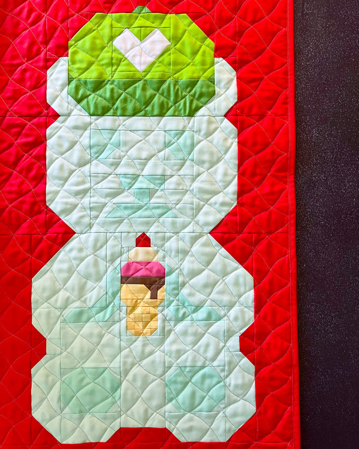 A close-up of a quilt featuring a bear design. The bear has a light aqua body, a green hat with a white heart, and holds a small ice cream cone.