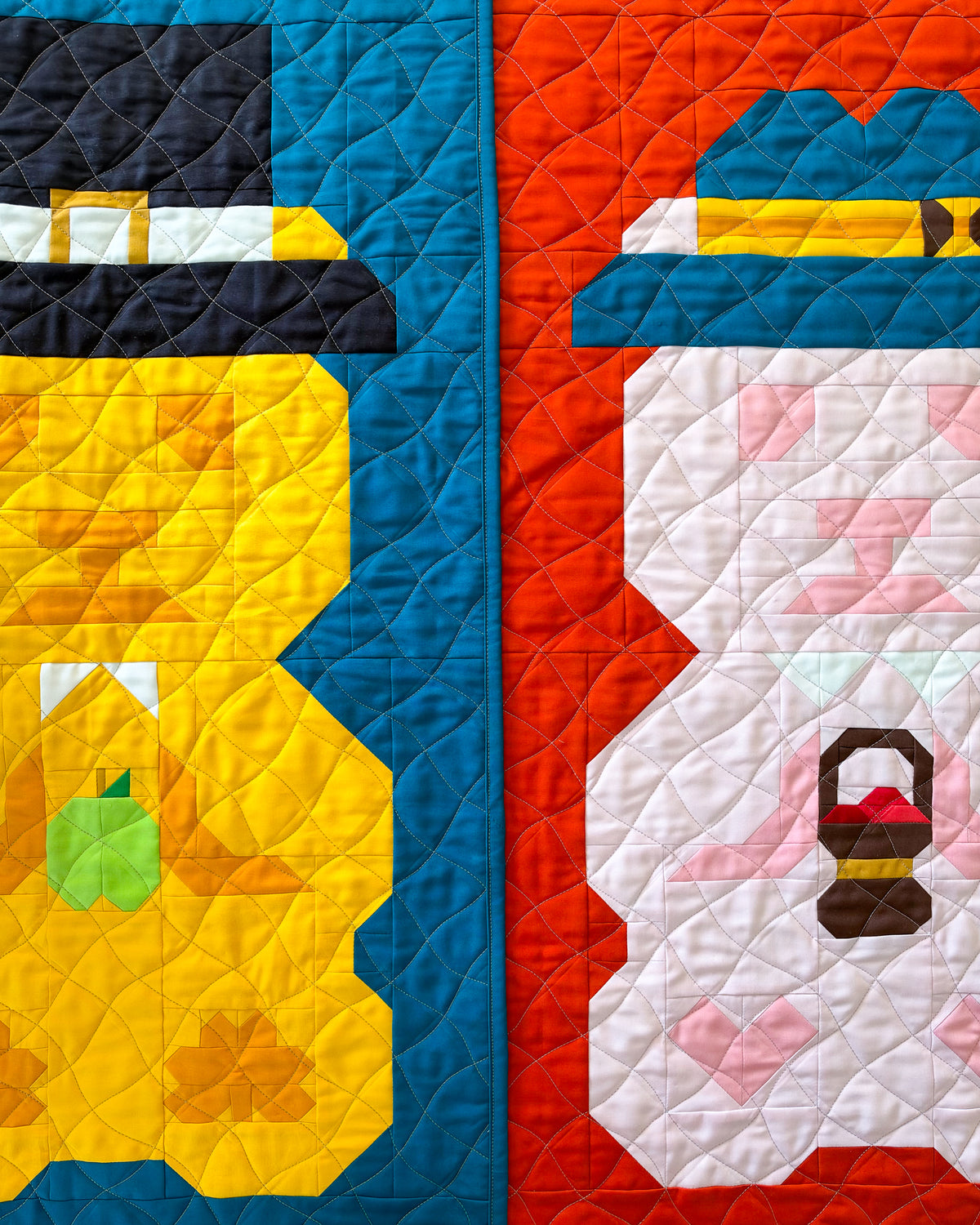 A close-up of a quilted textile featuring two stylized autumn themed bear figures side by side. The left figure is yellow with a black hat, white trim, and a green apple detail, bordered by a blue quilted section.