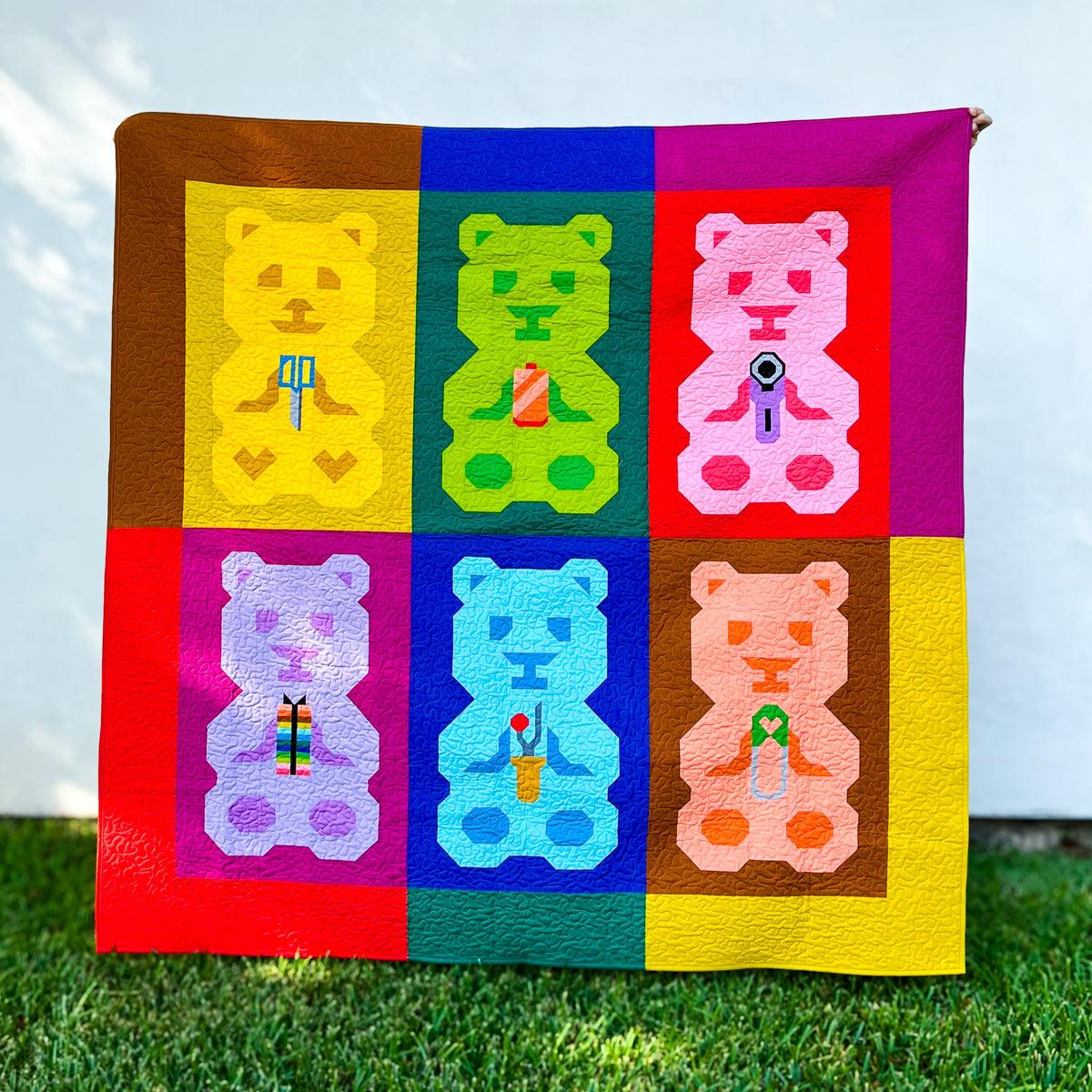 A colorful handmade quilt featuring a 2x3 grid of colorful teddy bears in bright yellow, green, pink, purple, blue, and peach tones, bordered by a matching multicolor patchwork design. The quilt is displayed outdoors on green grass against a white wall with soft leaf shadows. The quilt pattern is 'Quilty Beary Bunch'.