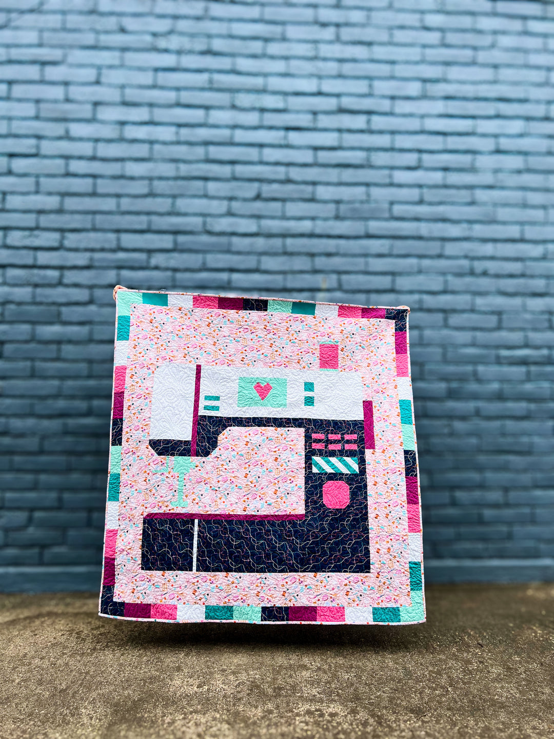 Oh The Possibilities quilt with sewing machine design against a brick wall.